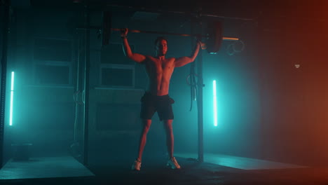 Male-powerlifter-in-a-blue-orange-neon-light-is-preparing-for-a-workout-in-the-gym.-A-strong-man-with-a-naked-torso-does-lifting-a-barbell-over-his-head-a-jerk-exercise-from-weightlifting.